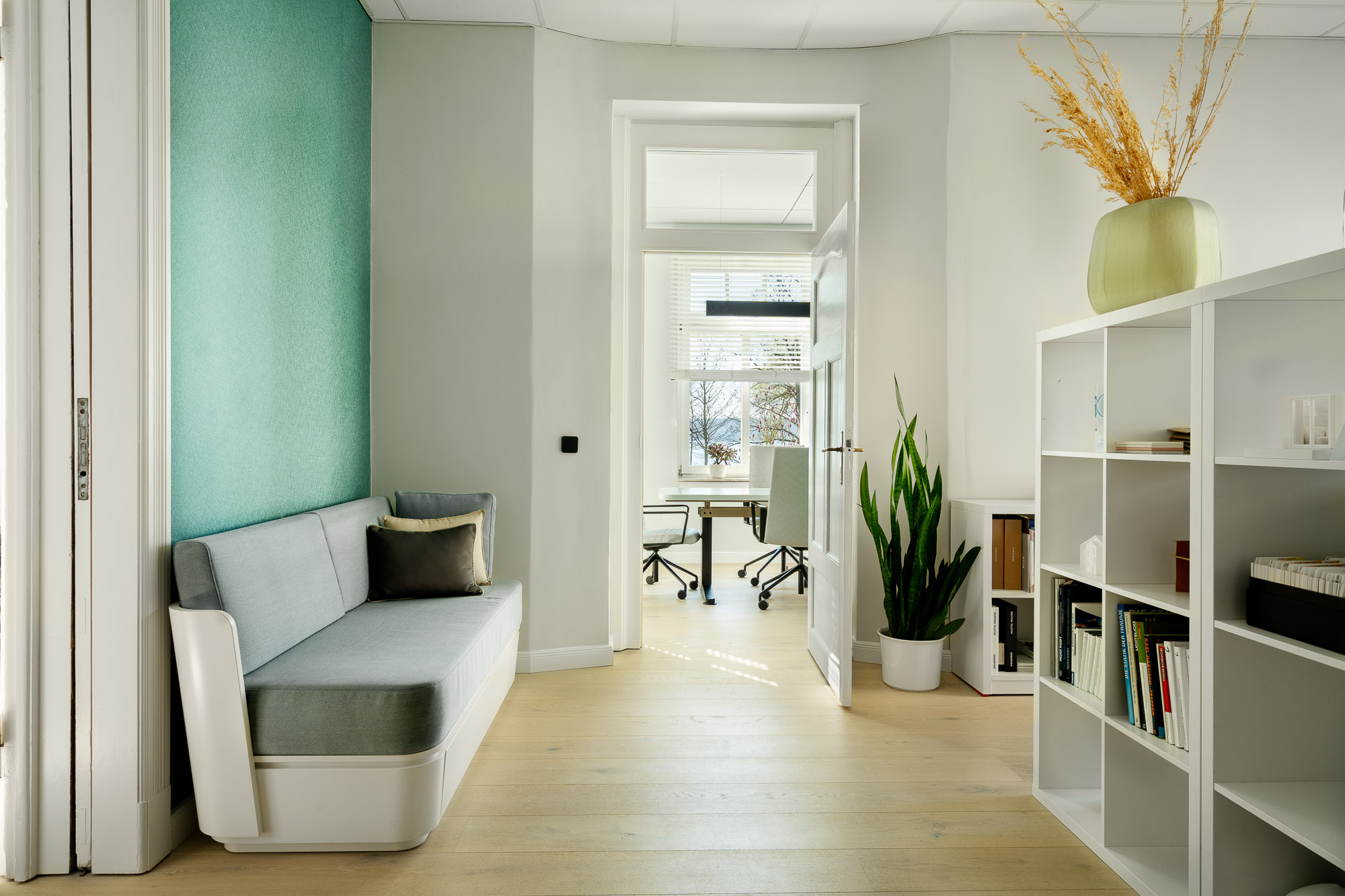 Modern office interior with a gray sofa, open door leading to a room with office chairs by a window, white shelving unit, and potted plants.